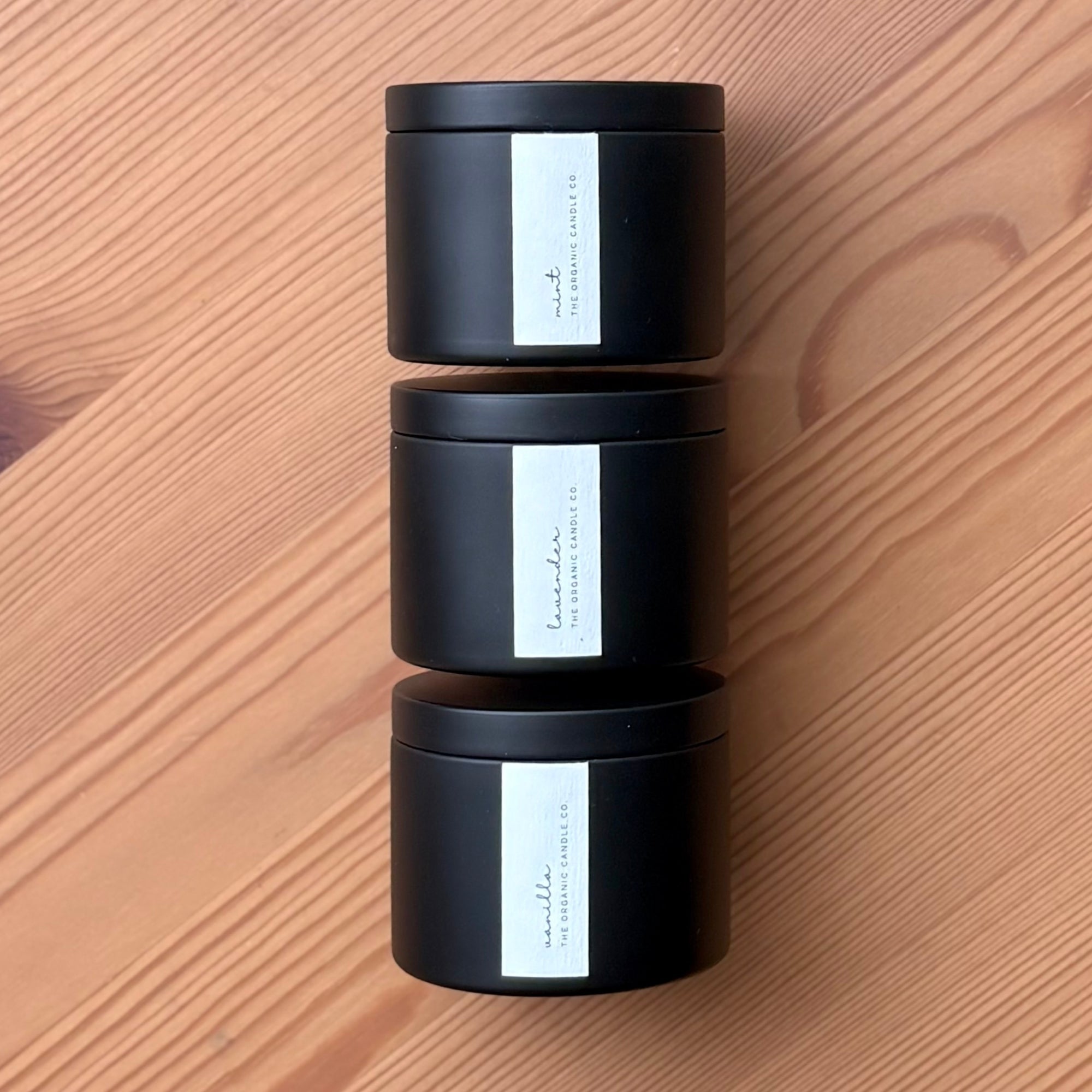 Three black organic candles with white labels stacked on a wooden surface