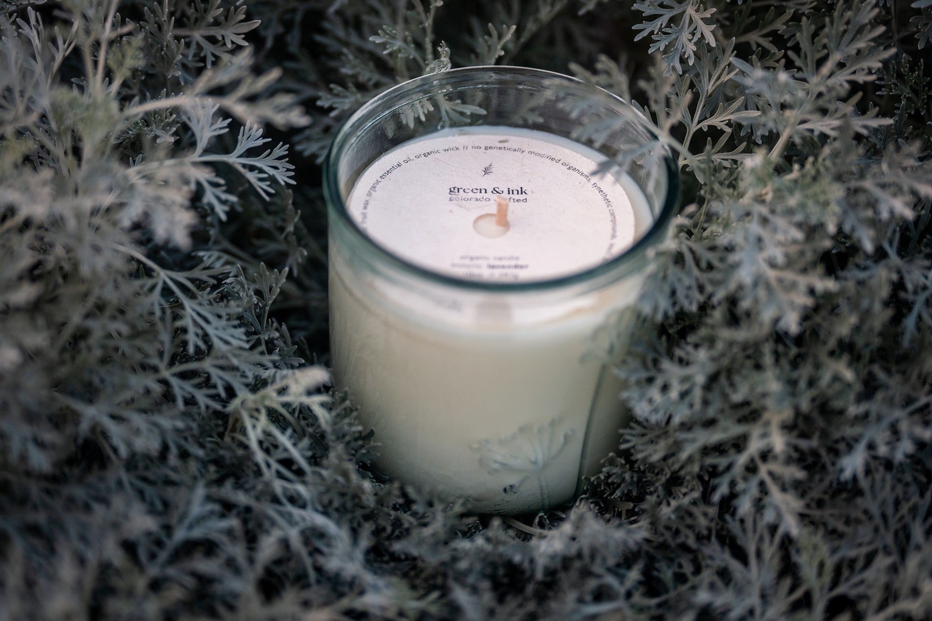 Organic candle in a recycled glass jar with a label on a sage-green bed of leaves