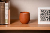 Organic candle in terracotta ceramic fair trade vessel on a wooden shelf with books and a framed print in the background