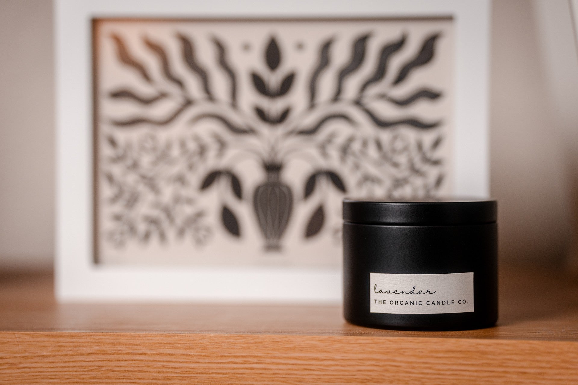 Black candle labeled 'Lavender' by 'The Organic Candle Co.' on a wooden surface with a decorative wall piece in the background.