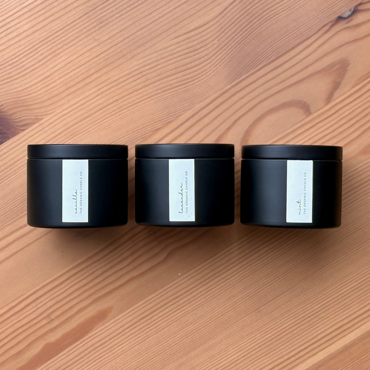 Three organic candles in black metal containers with labels on a wooden surface