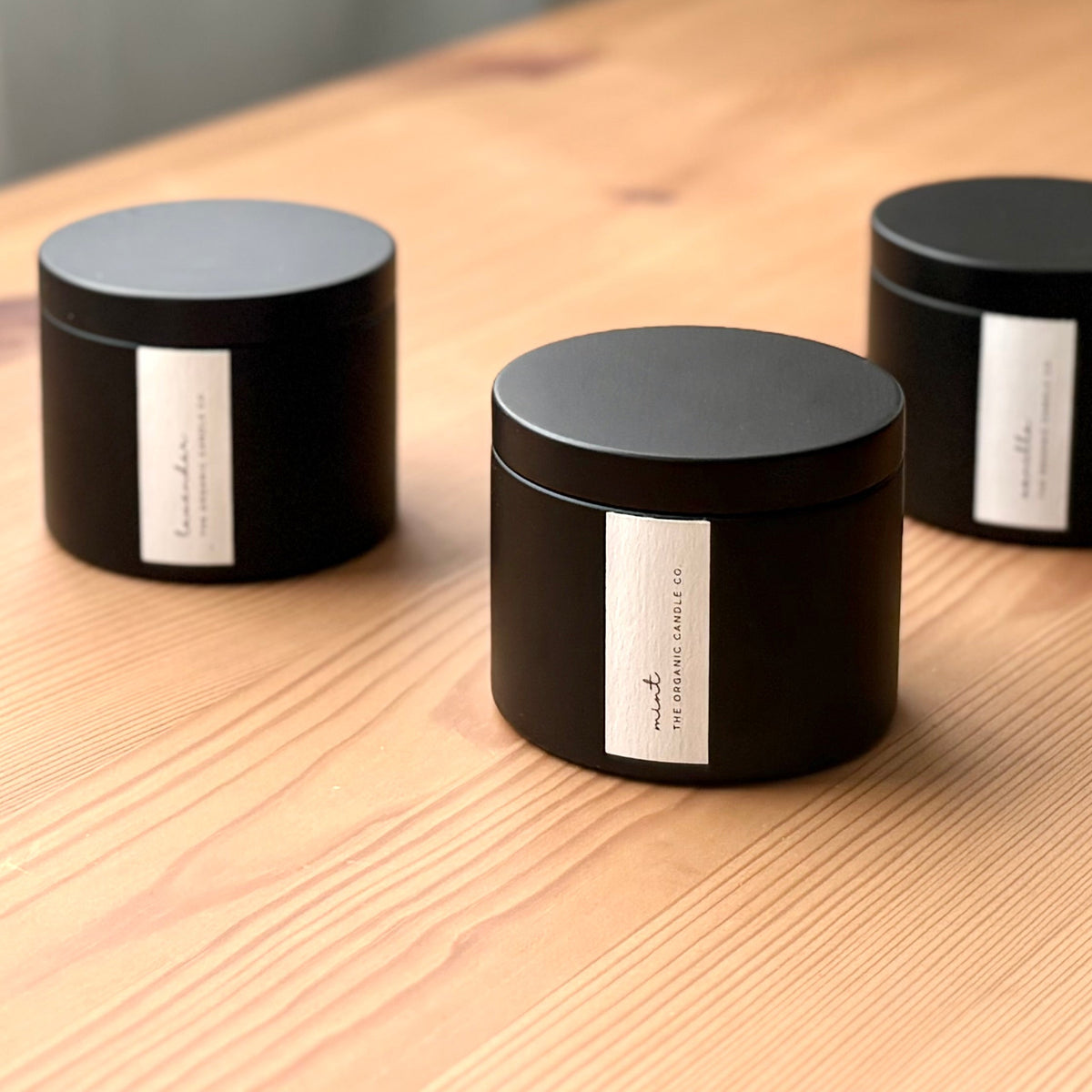 Three organic candles in black metal containers with labels on a wooden surface