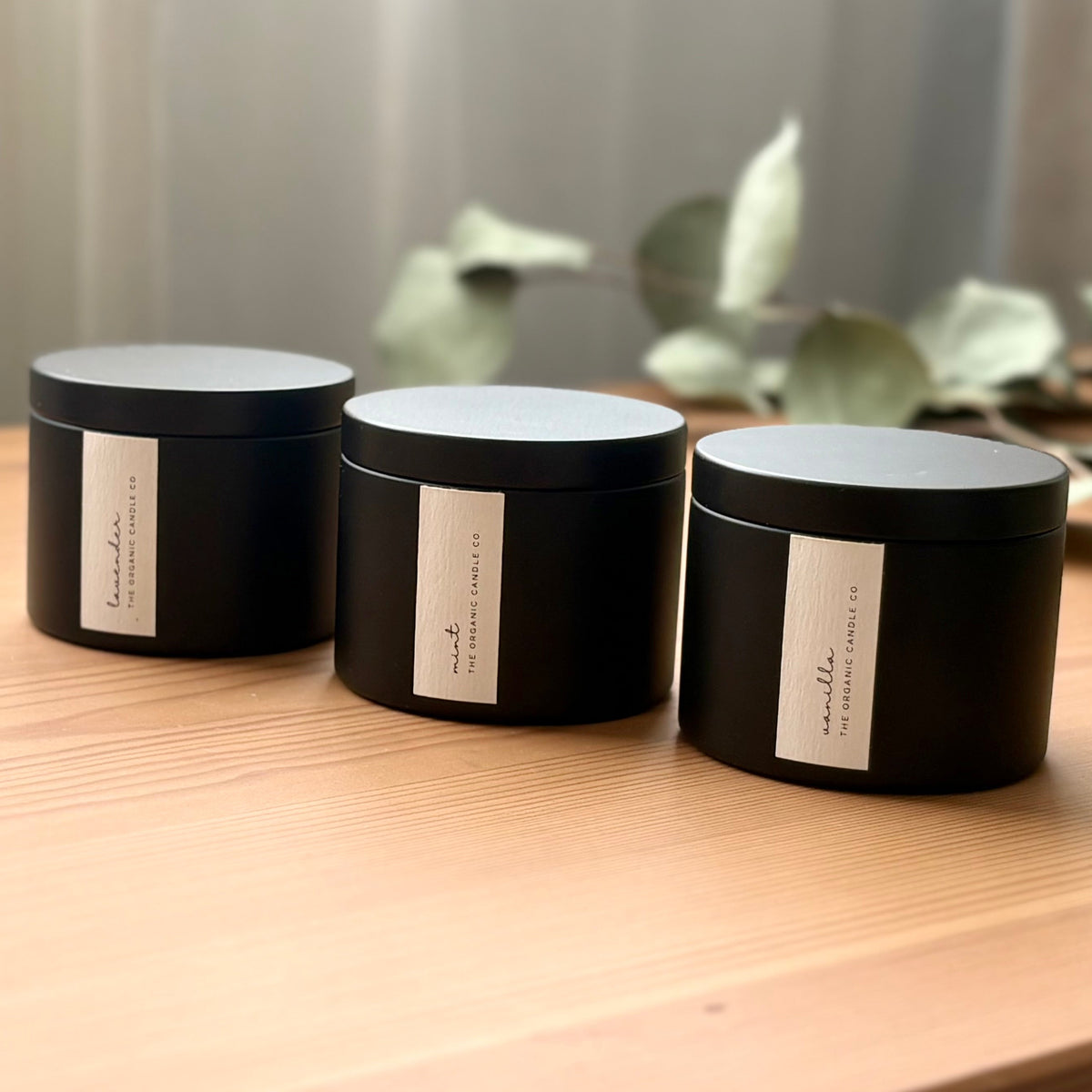 Three organic candles in black metal containers with labels on a wooden surface