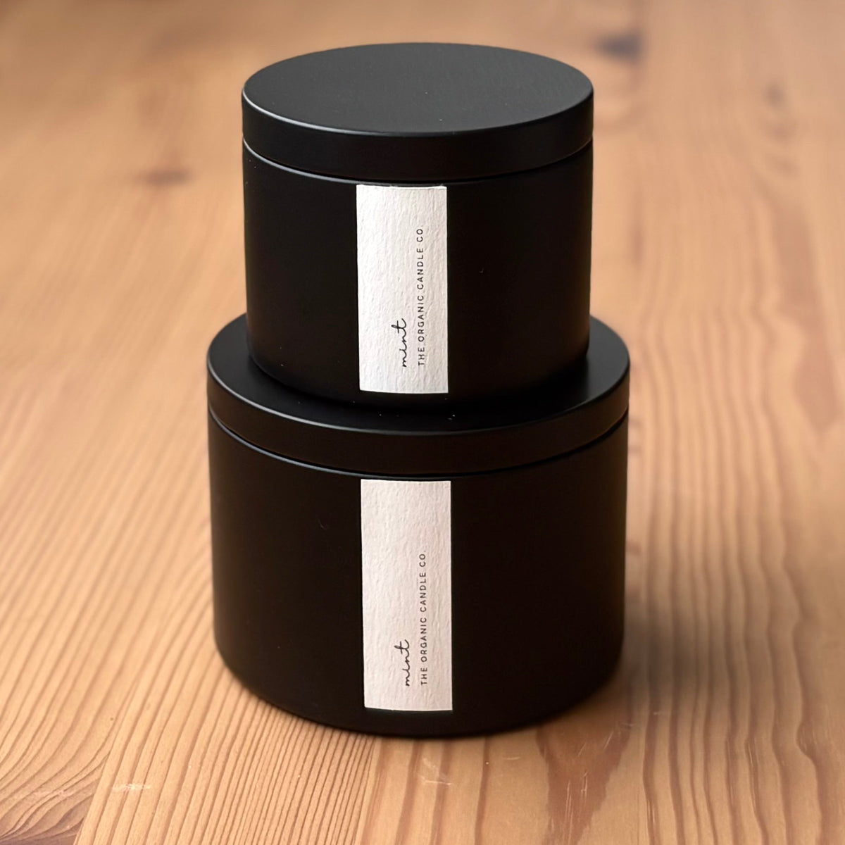 Two organic candles in black metal containers with labels on a wooden surface
