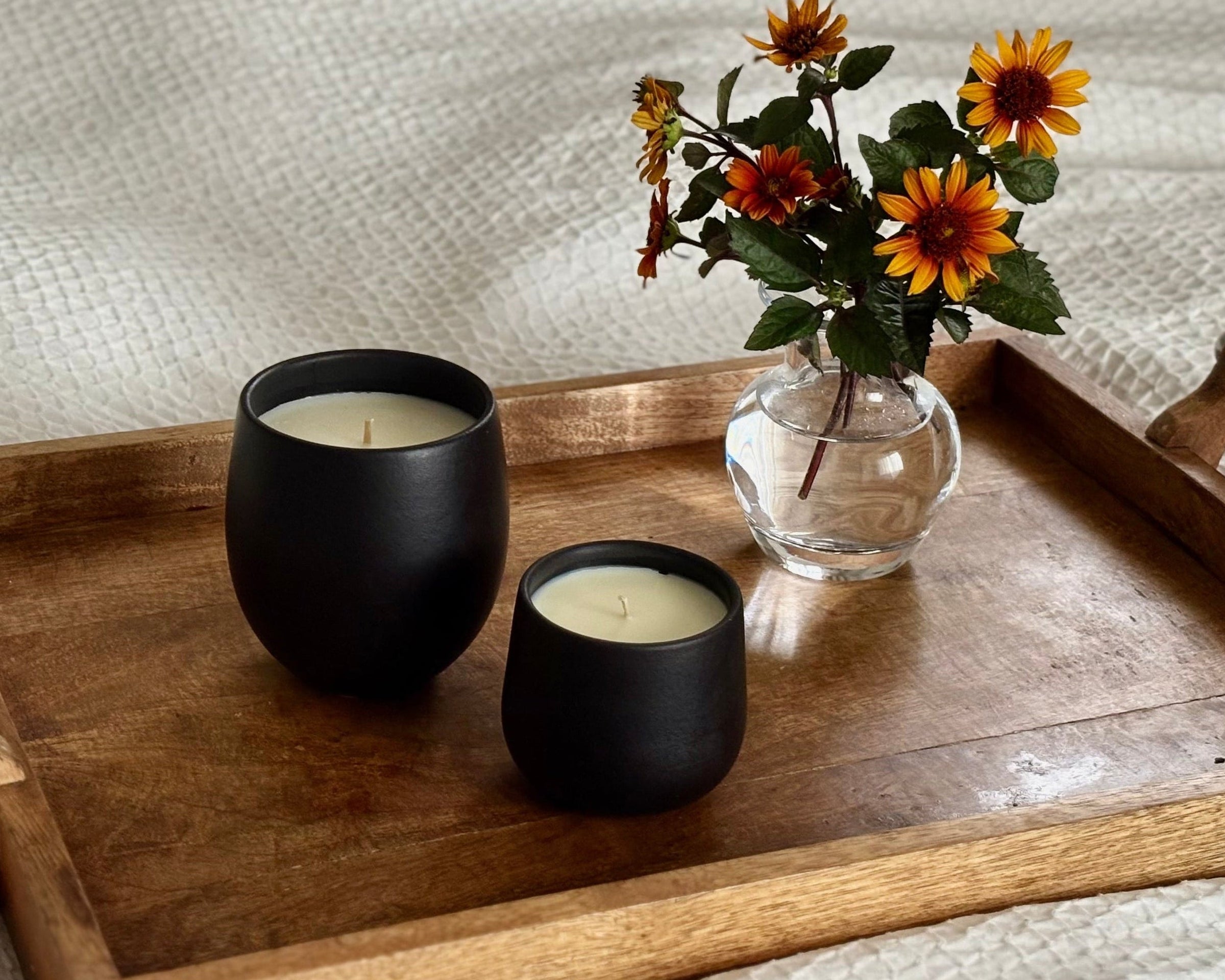 A wooden serving tray with metal handles rests on a white quilted bedspread. On the tray are two matte black organic candles and a small clear glass vase holding orange and yellow flowers.