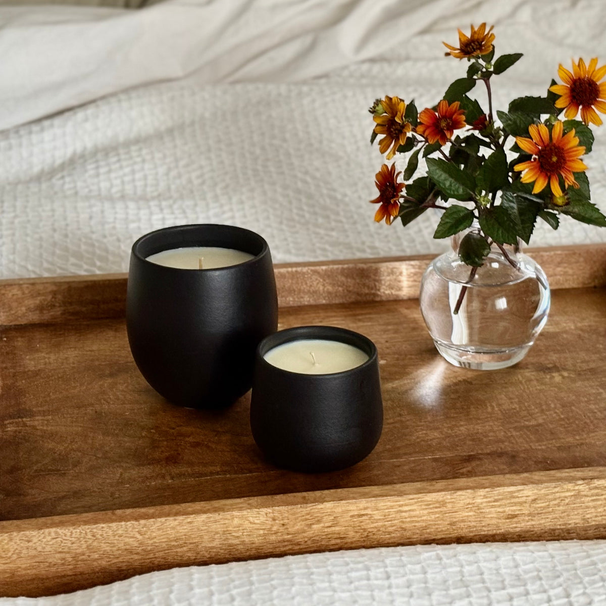 Two matte black ceramic organic candles on a wooden tray beside a small glass vase with orange flowers, set on a bed with white textured bedding.