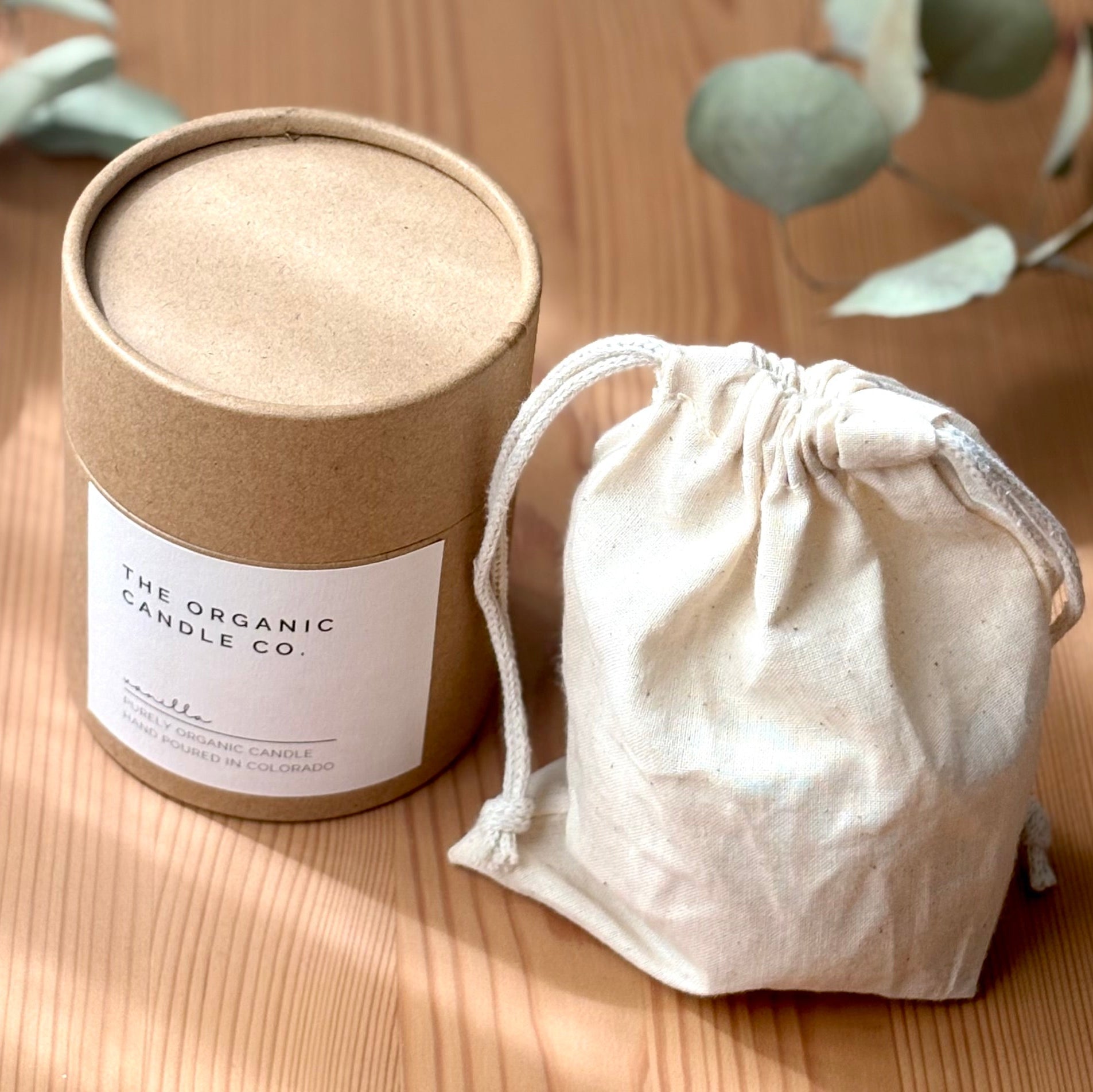 Cylindrical container with a "The Organic Candle Co." label and an organic muslin bag on a wooden surface with eucalyptus leaves.