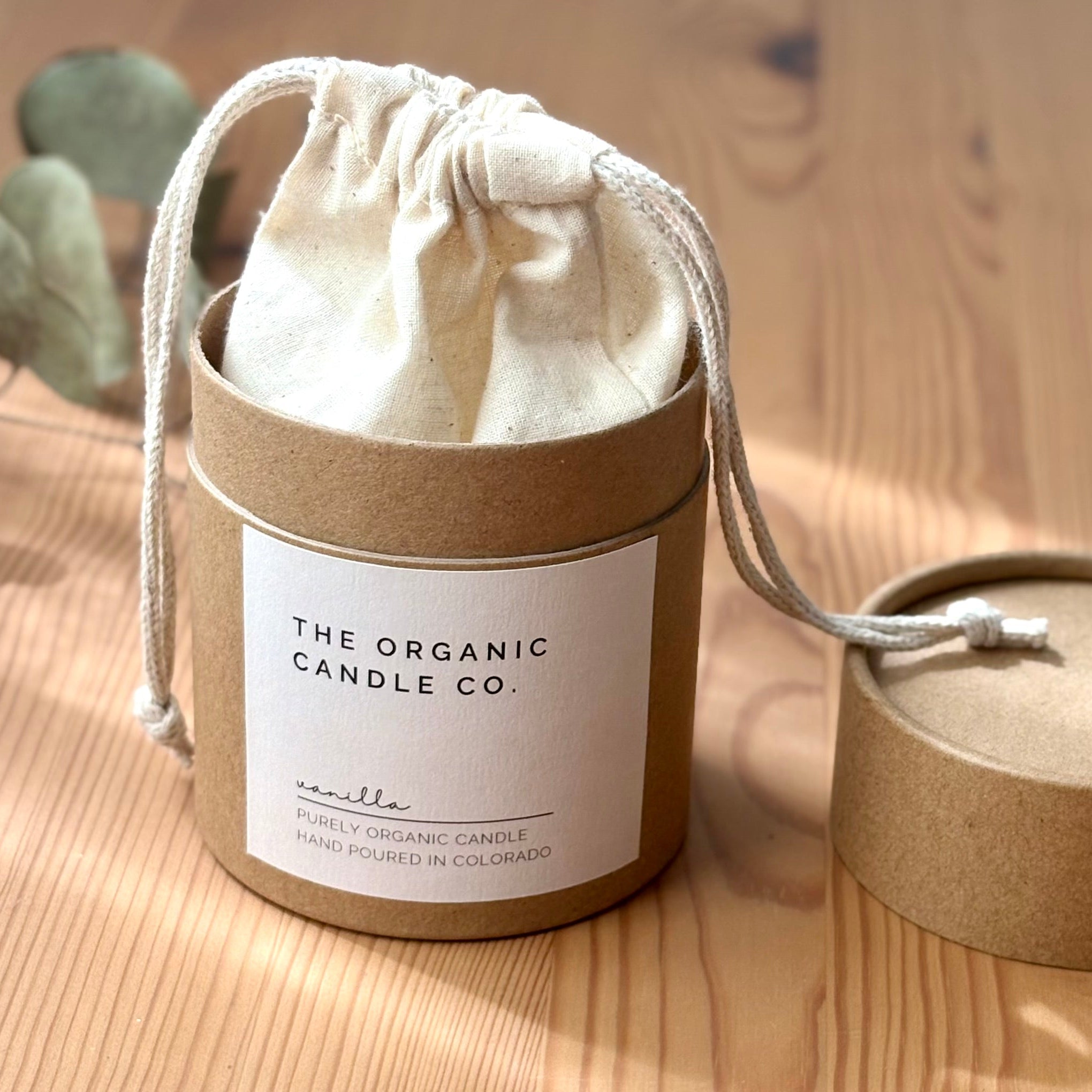 Candle in a cardboard container with a drawstring lid on a wooden surface, featuring &