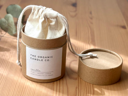 Candle in a cardboard container with a drawstring lid on a wooden surface, featuring &