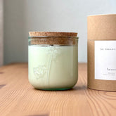 Organic candle in a recycled glass floral jar with a kraft cylinder box on a wooden surface. A white label on the box reads: "The Organic Candle Co.; lavender"