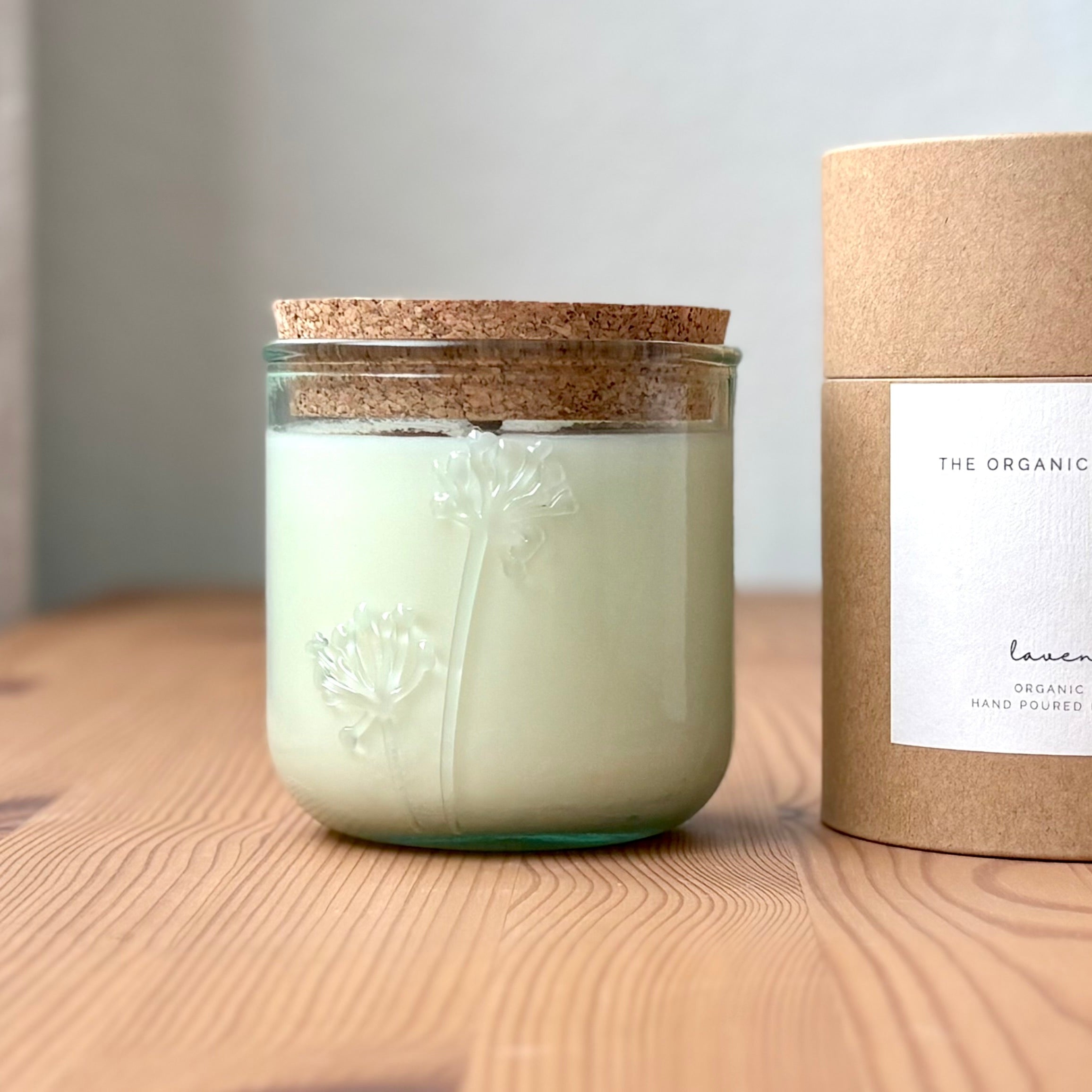 Organic candle in a recycled glass floral jar with a kraft cylinder box on a wooden surface. A white label on the box reads: "The Organic Candle Co.; lavender"