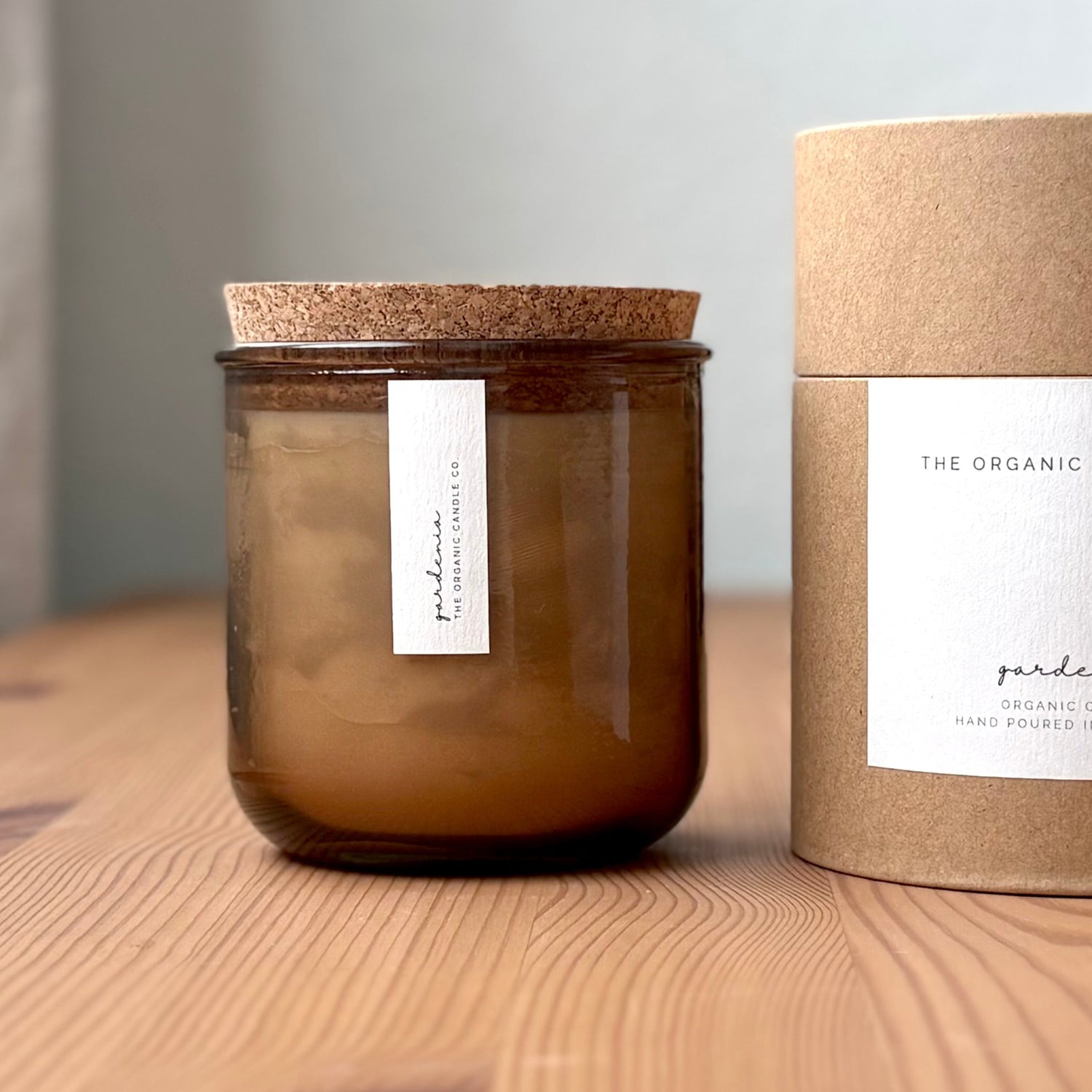 Organic candle in rose recycled glass jar on a wooden background, with a cork top. A label reads "gardenia; The Organic Candle Co." Next to it is a cylinder kraft packaging with "The Organic Candle Co." label.