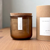 Organic candle in rose recycled glass jar on a wooden background, with a cork top. A label reads "gardenia; The Organic Candle Co." Next to it is a cylinder kraft packaging with "The Organic Candle Co." label.