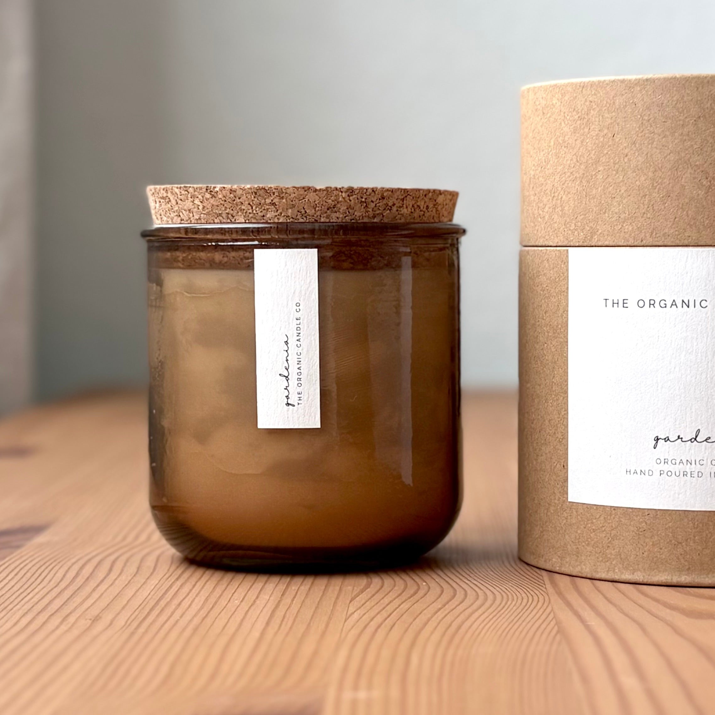 Organic candle in rose recycled glass jar on a wooden background, with a cork top. A label reads "gardenia; The Organic Candle Co." Next to it is a cylinder kraft packaging with "The Organic Candle Co." label.