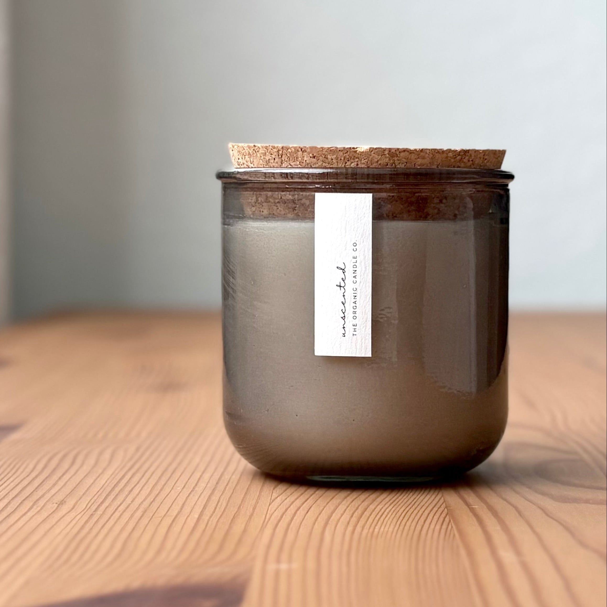 Organic candle in light gray recycled glass jar on a wooden background, with a cork top. A label reads "unscented; The Organic Candle Co."