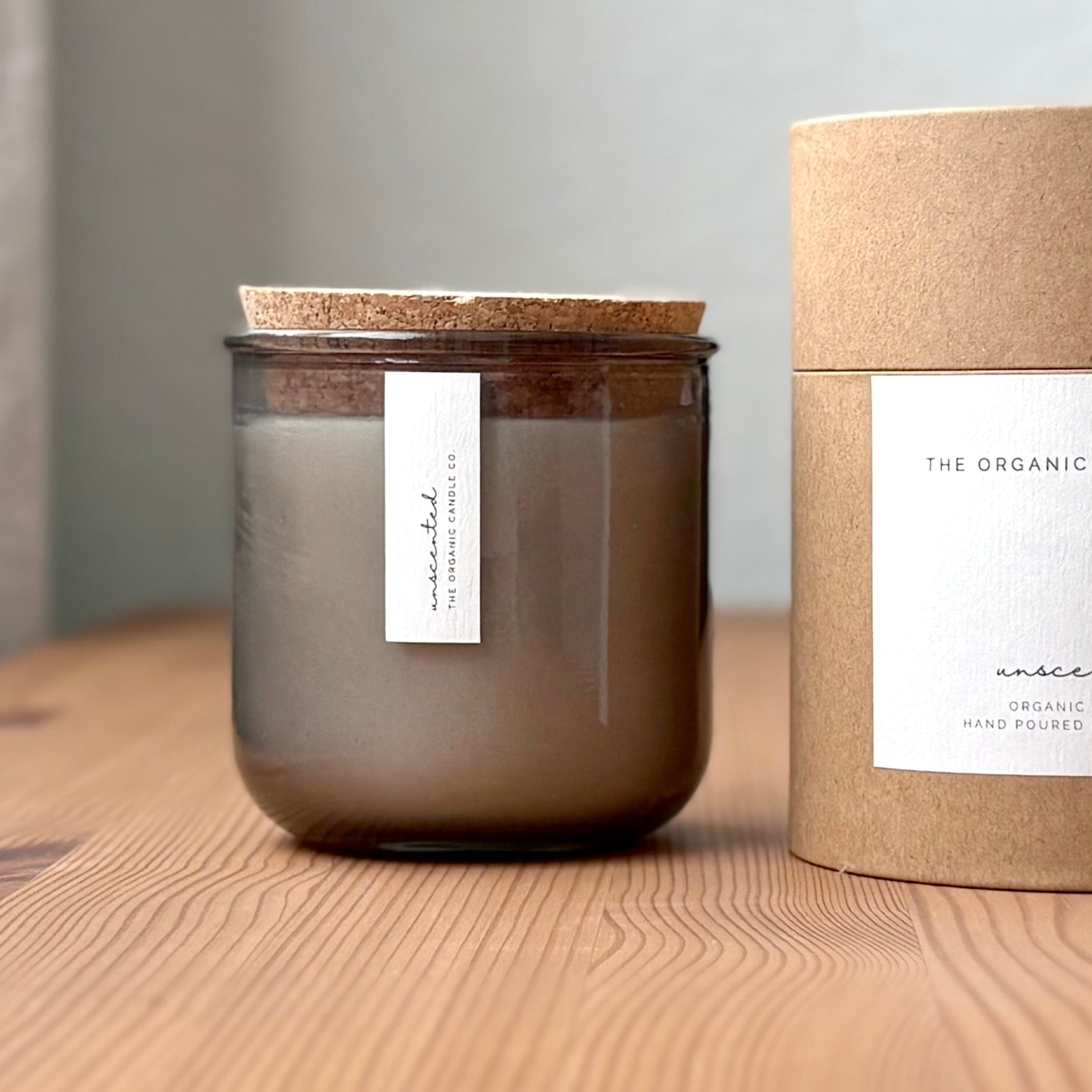 Organic candle in light gray recycled glass jar on a wooden background, with a cork top. A label reads "unscented; The Organic Candle Co." Next to it is a cylinder kraft packaging with "The Organic Candle Co." label.