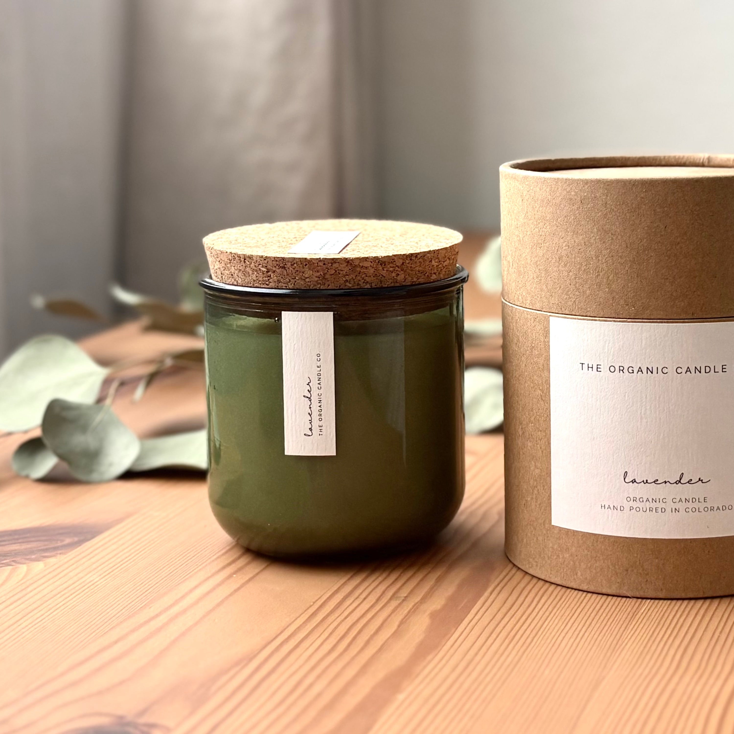 Organic candle in green recycled glass jar on a wooden background. A label reads "lavender; The Organic Candle Co." Next to it is a cylinder kraft packaging with "The Organic Candle Co." label.