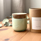 Organic candle in a recycled glass floral jar with a kraft cylinder box on a wooden surface. A white label on the box reads: "The Organic Candle Co.; lavender"