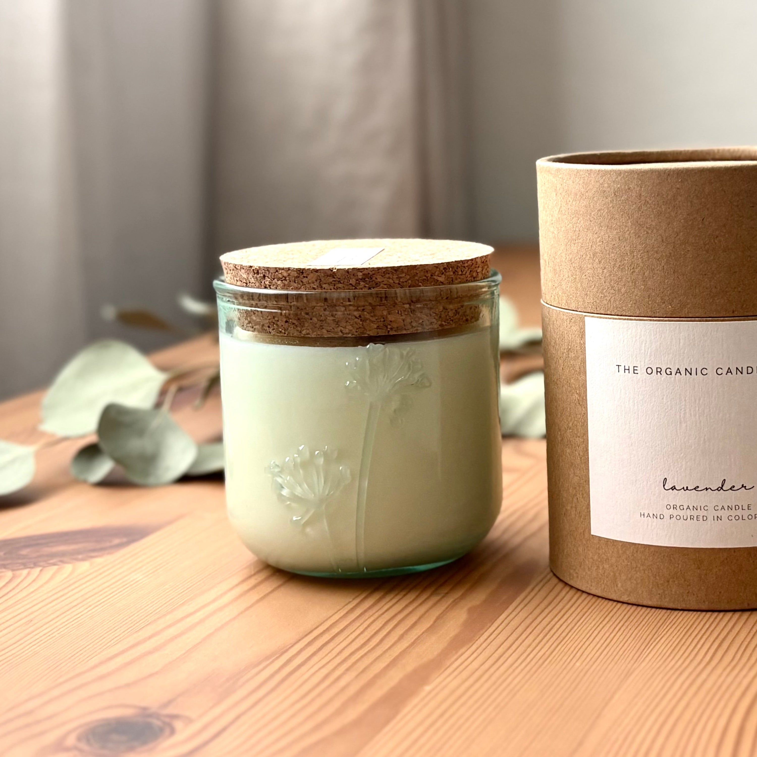 Organic candle in a recycled glass floral jar with a kraft cylinder box on a wooden surface. A white label on the box reads: "The Organic Candle Co.; lavender"