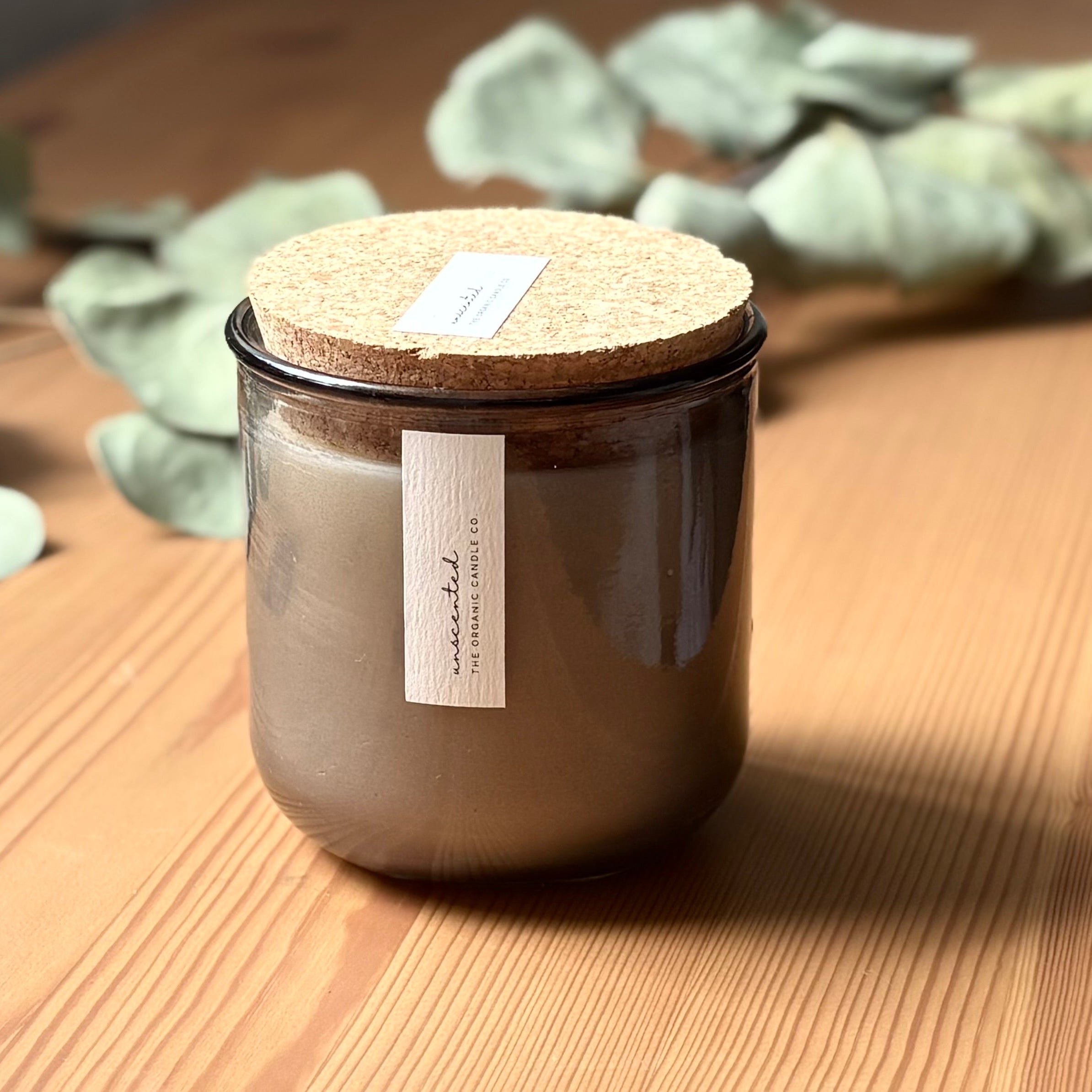 Organic candle in light gray recycled glass jar on a wooden background, with a cork top. A label reads "unscented; The Organic Candle Co."