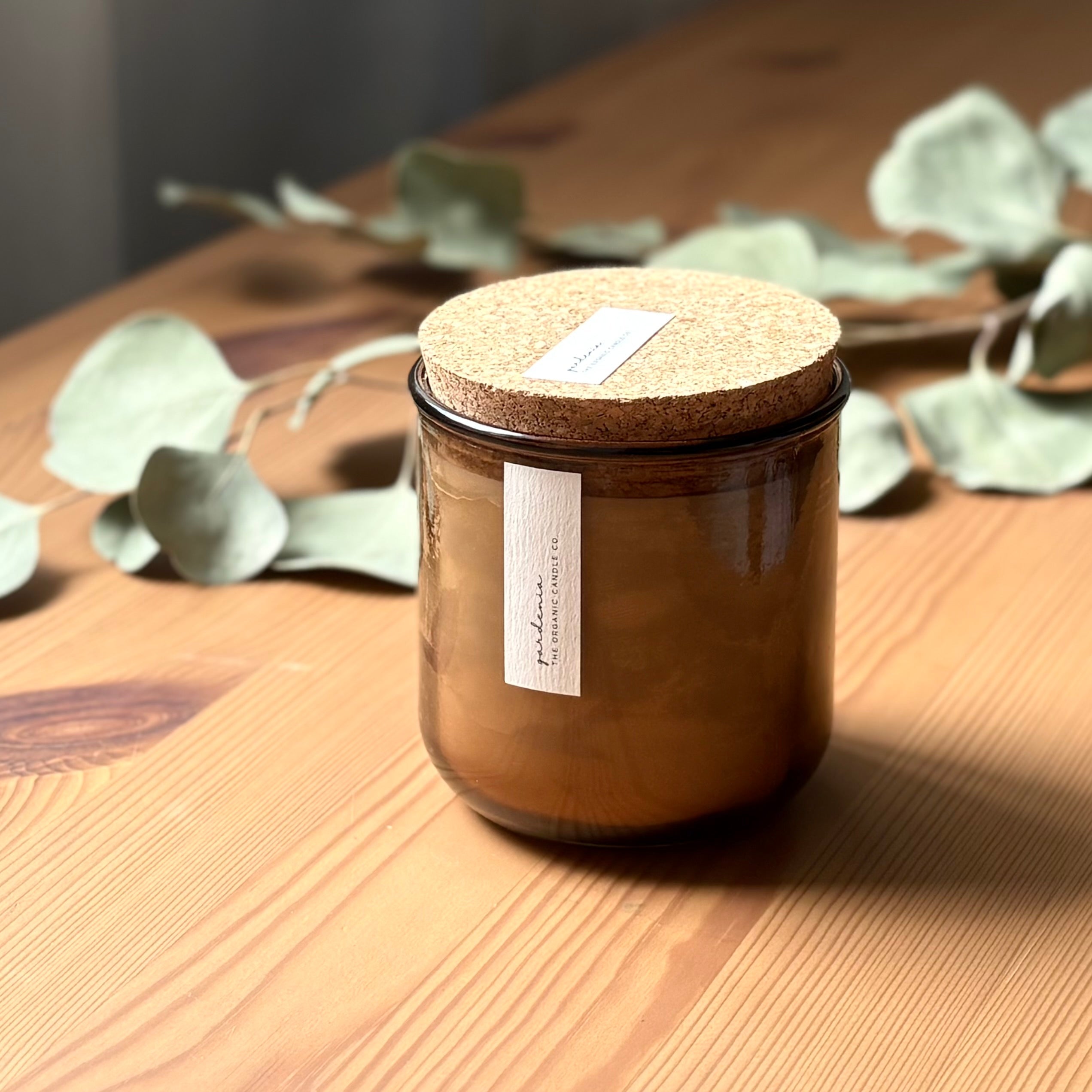 Organic candle in rose recycled glass jar on a wooden background, with a cork top. A label reads "gardenia; The Organic Candle Co."