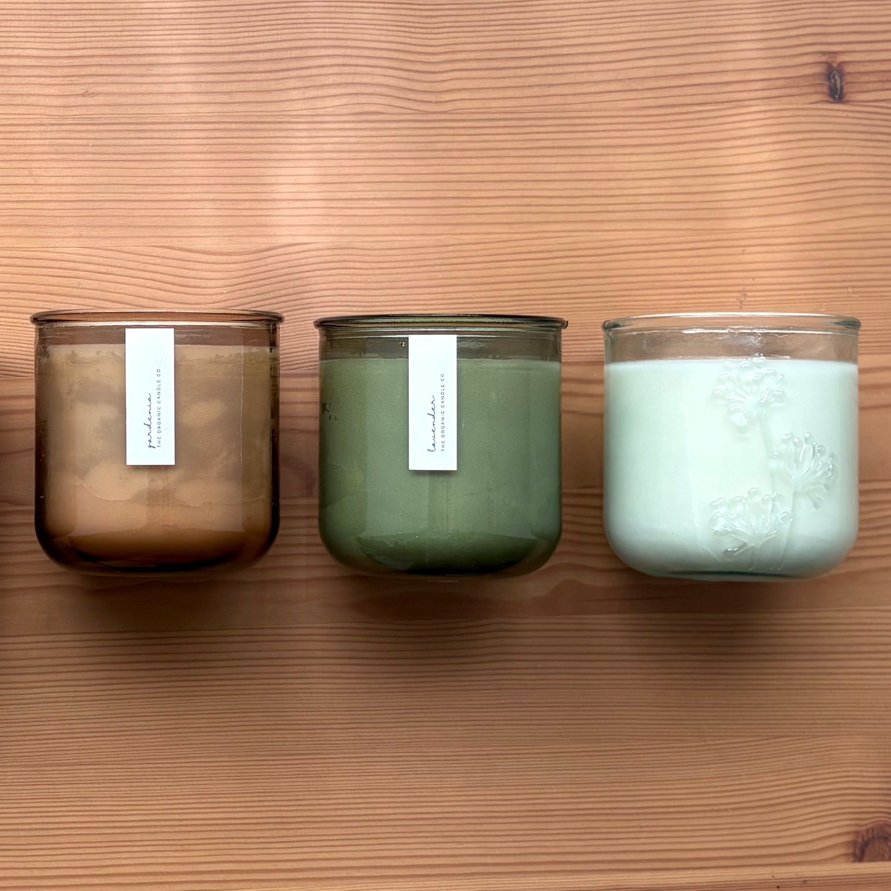 Organic candles in various colors of recycled glass jars on a wooden background. Thin white labels read: "The Organic Candle Co."
