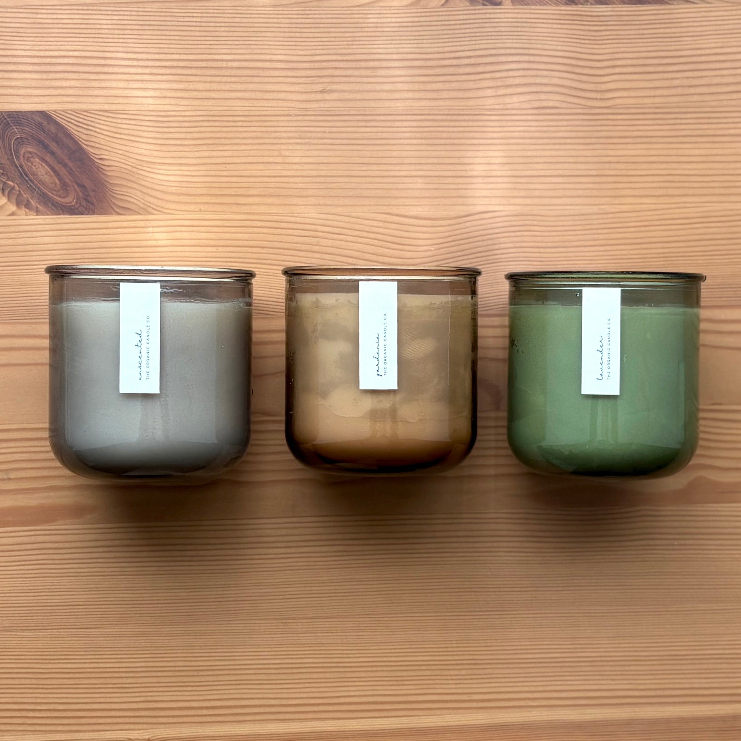 Organic candles in various colors of recycled glass jars on a wooden background. Thin white labels read: "The Organic Candle Co."