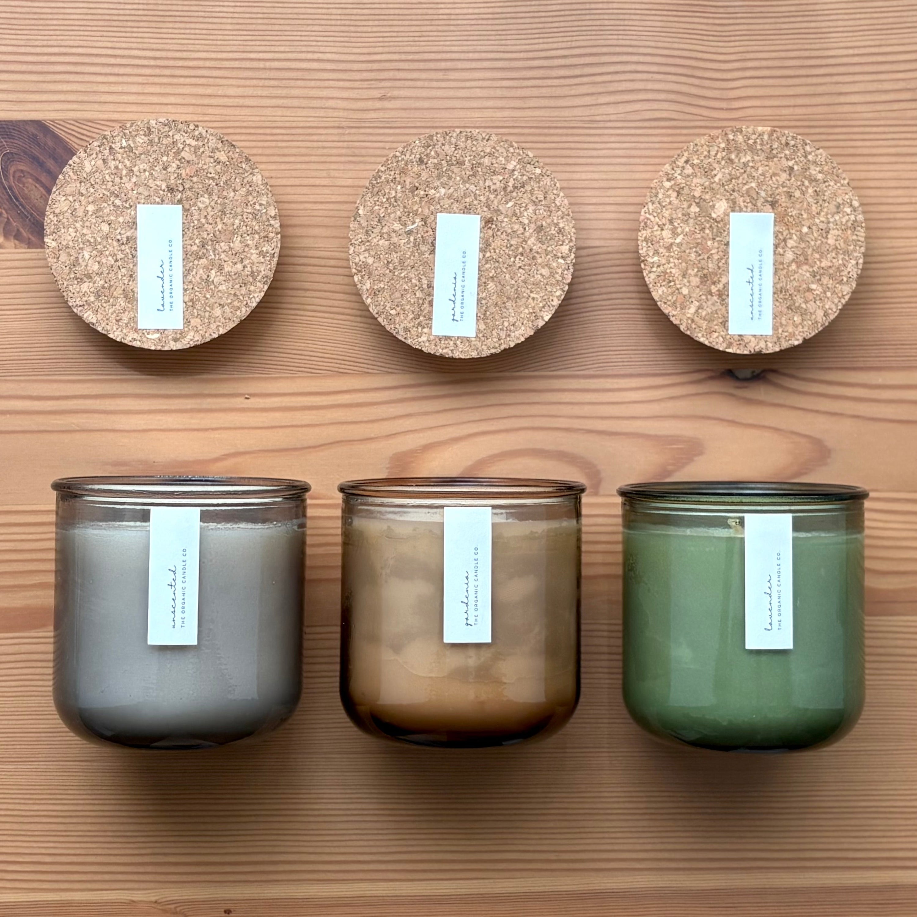 Organic candles in various colors of recycled glass jars on a wooden background, with cork tops laying above. Thin white labels read: "The Organic Candle Co."