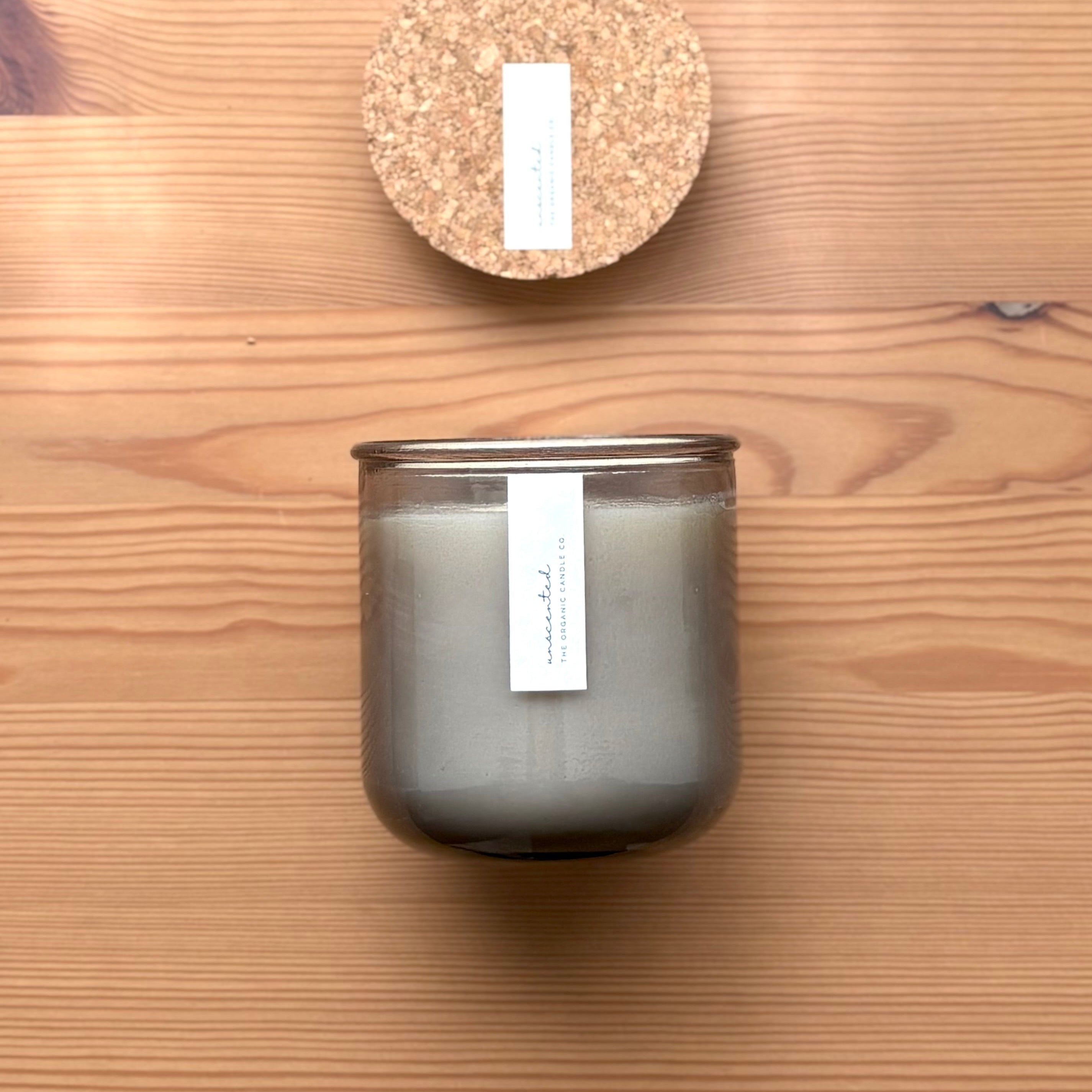 Organic candle in light gray recycled glass jar on a wooden background, with a cork top above. A label reads "unscented; The Organic Candle Co."