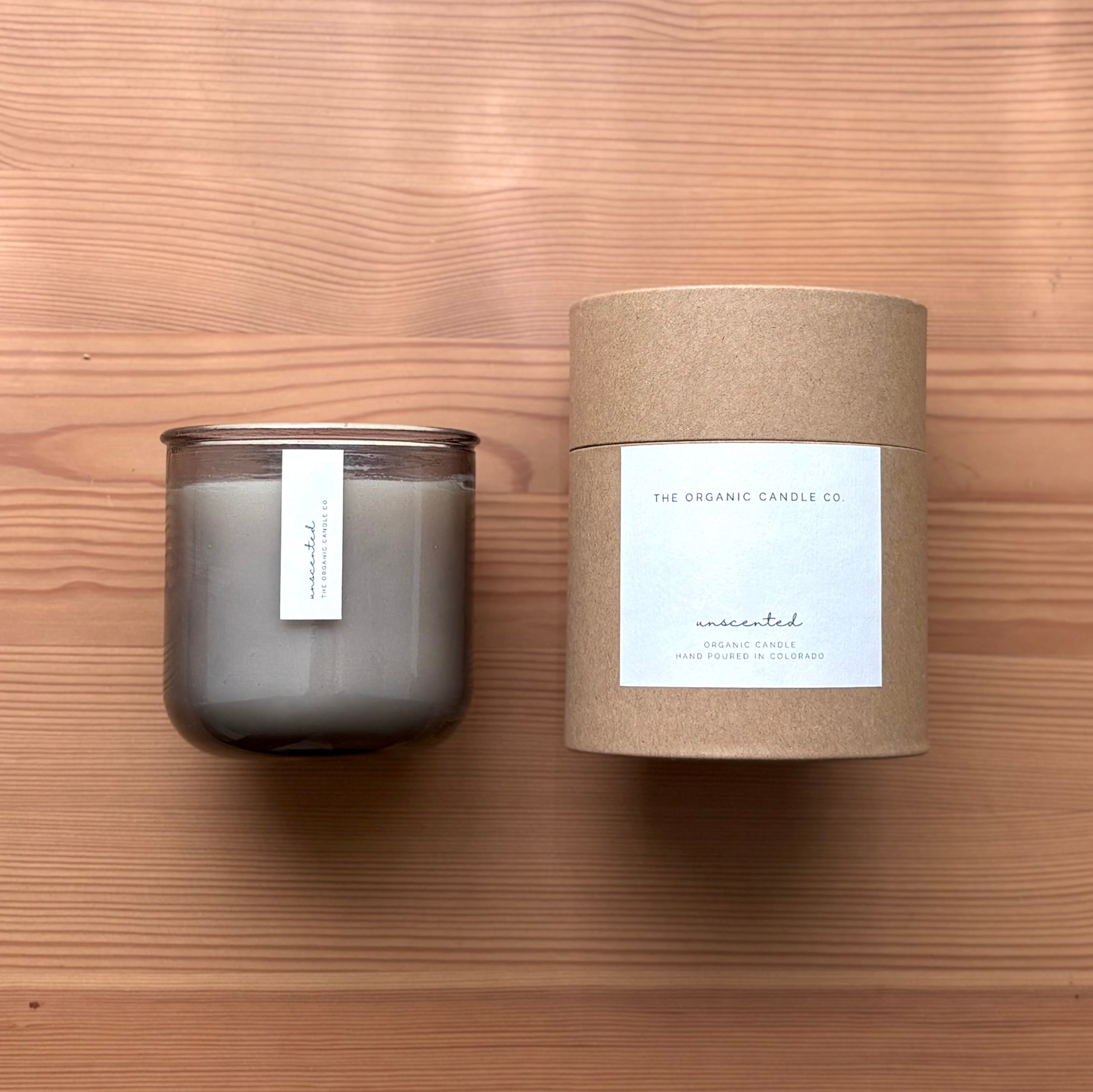 Organic candle in light gray recycled glass jar on a wooden background. A label reads "unscented; The Organic Candle Co." Next to it is a cylinder kraft packaging with "The Organic Candle Co." label.