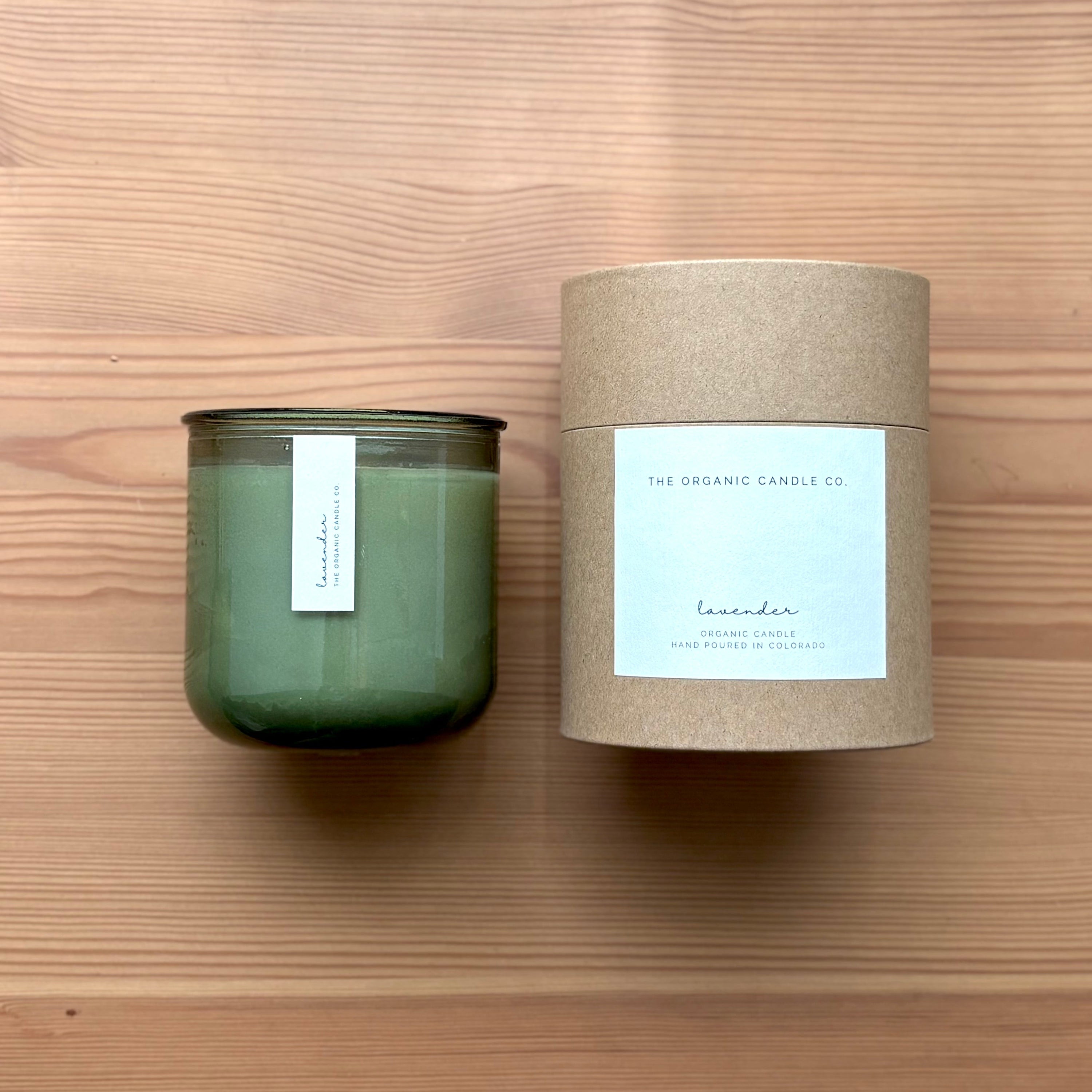 Organic candle in green recycled glass jar on a wooden background. A label reads "lavender; The Organic Candle Co." Next to it is a cylinder kraft packaging with "The Organic Candle Co." label.