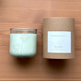 Organic candle in a recycled glass floral jar with a kraft cylinder box on a wooden surface. A white label on the box reads: "The Organic Candle Co.; lavender"