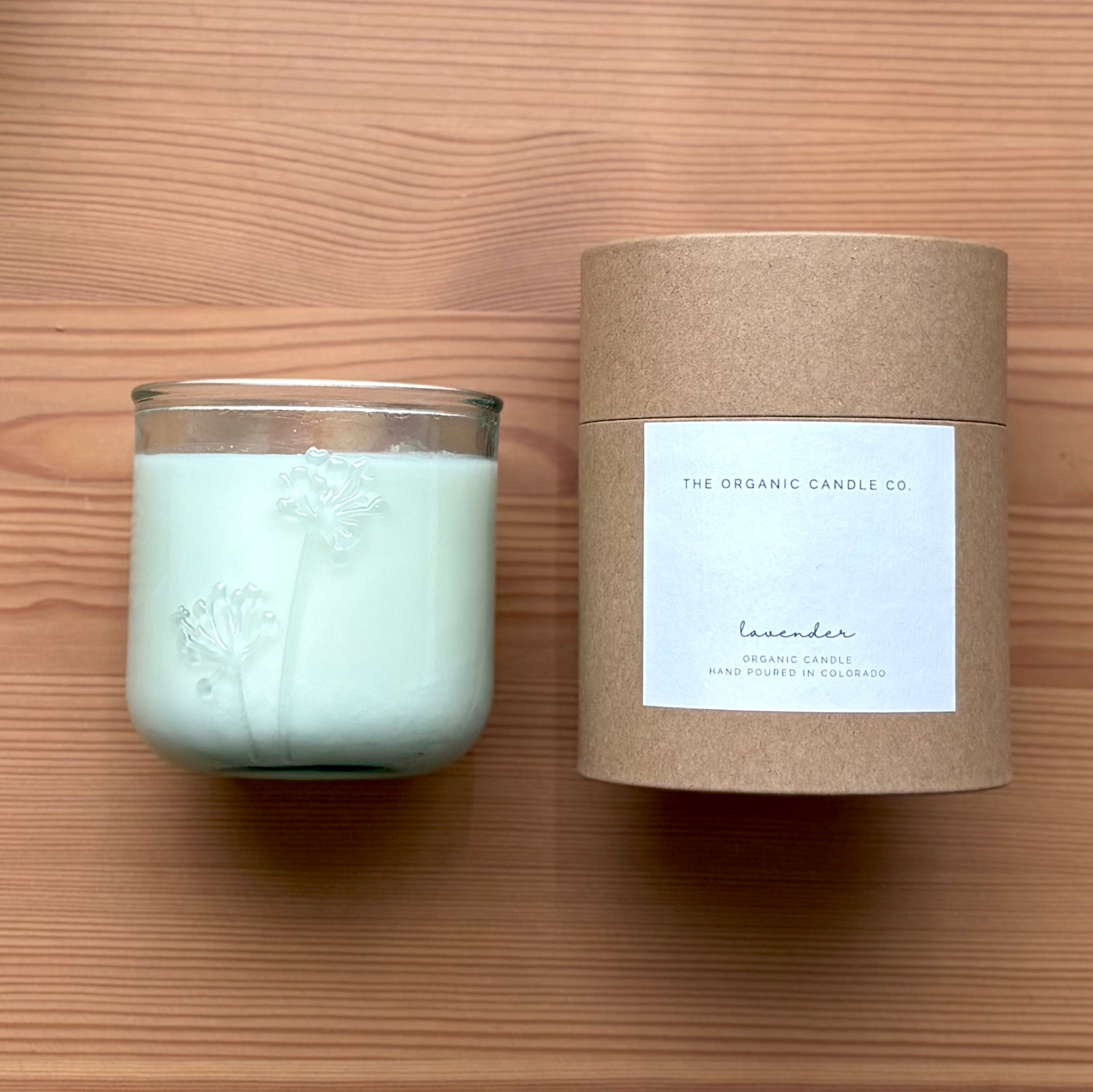 Organic candle in a recycled glass floral jar with a kraft cylinder box on a wooden surface. A white label on the box reads: "The Organic Candle Co.; lavender"
