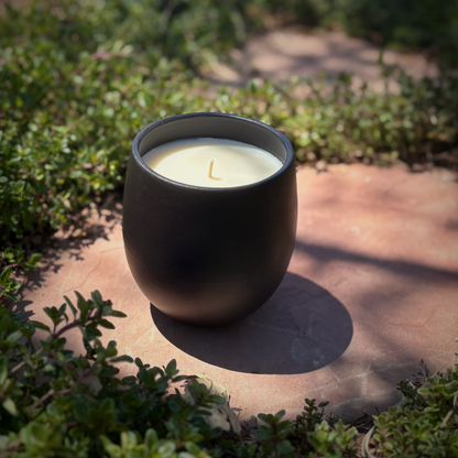 A matte black organic candle placed on a stone path outdoors with green foliage in the background.
