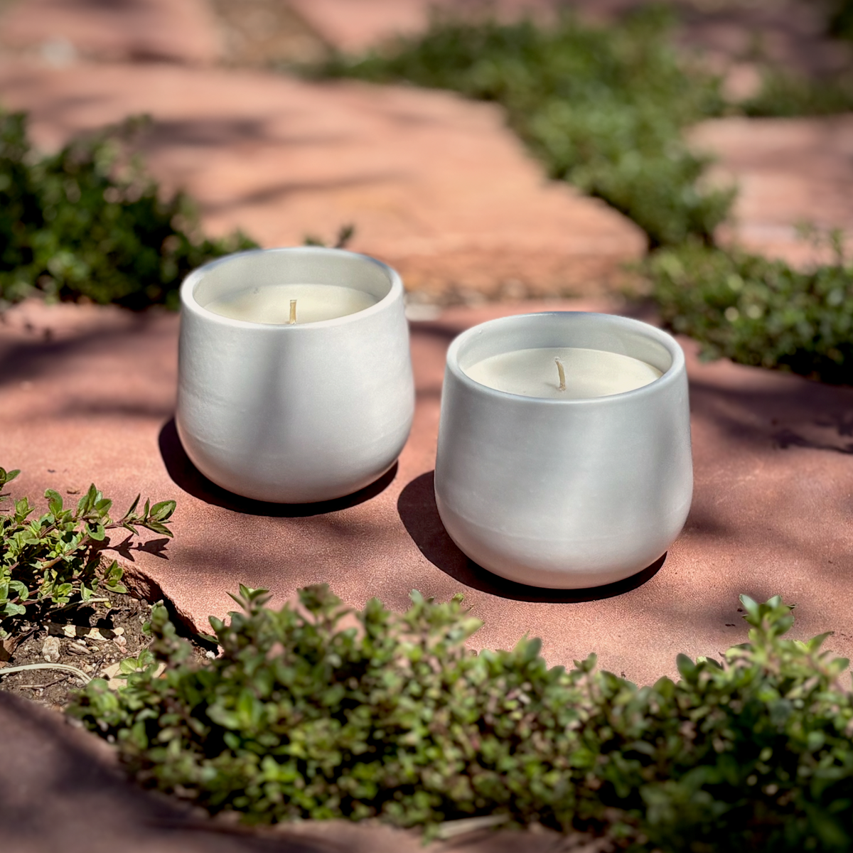 Two white ceramic organic candles in matching holders, placed on a stone path surrounded by green plants in an outdoor garden setting.