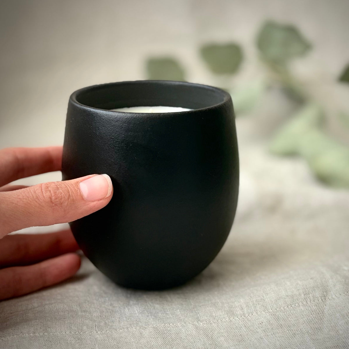 A matte black organic candle being held by a hand, set against a soft beige fabric backdrop with blurred green leaves in the background.