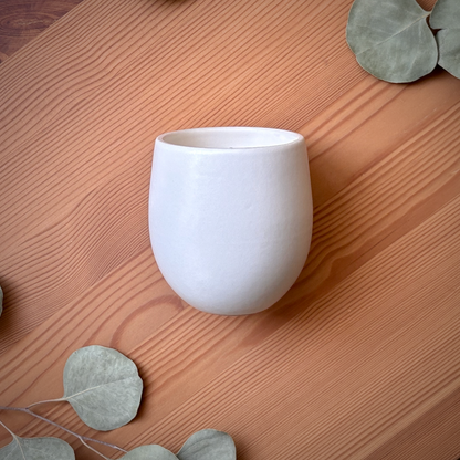 A matte white ceramic organic candle placed on a warm brown wooden surface with decorative eucalyptus leaves.
