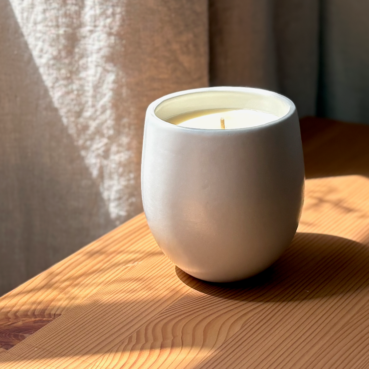 A minimalist matte white ceramic organic candle with a rounded, organic shape, resting on a warm wooden table in soft natural light.