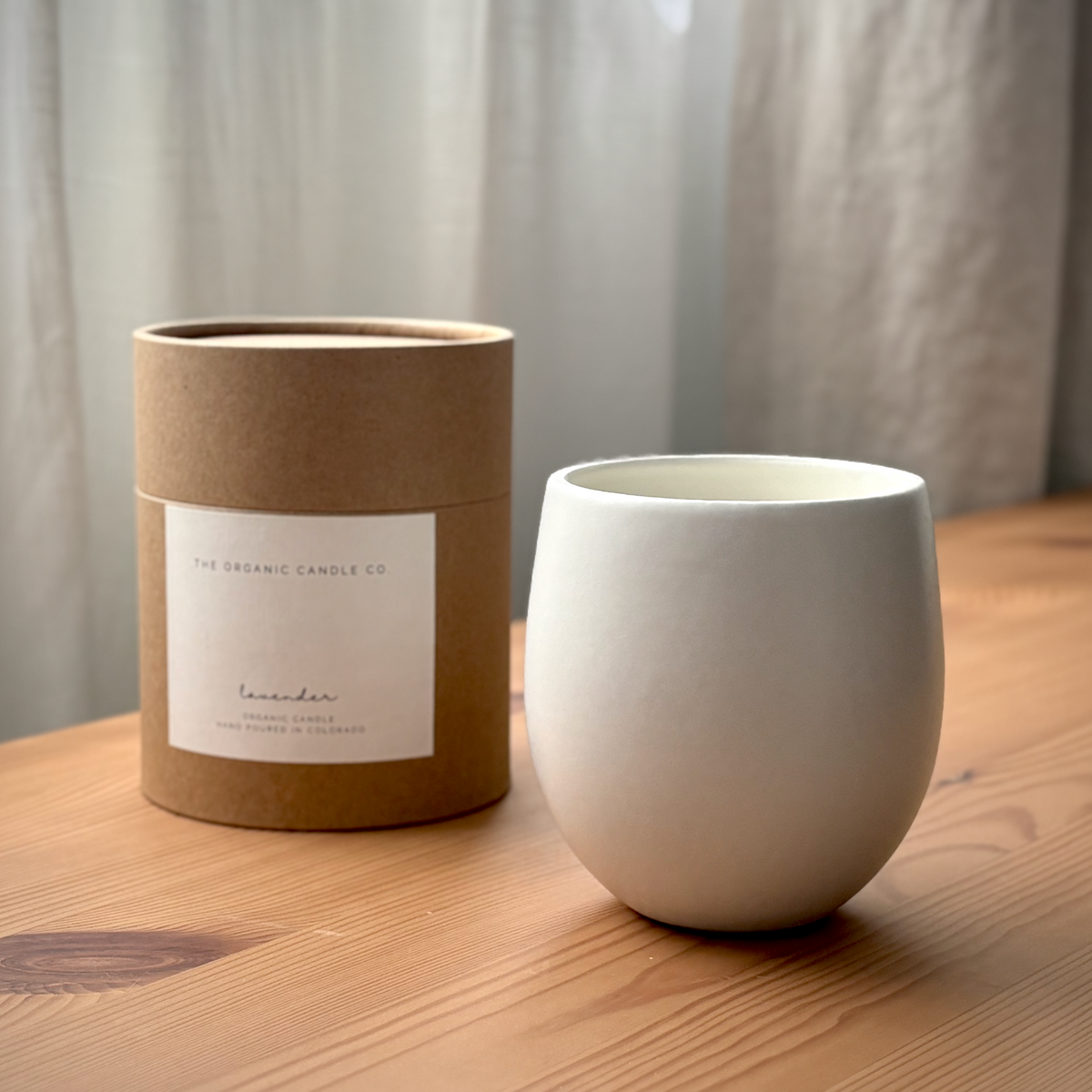 A white ceramic candle next to its brown kraft paper packaging cylinder, labeled &