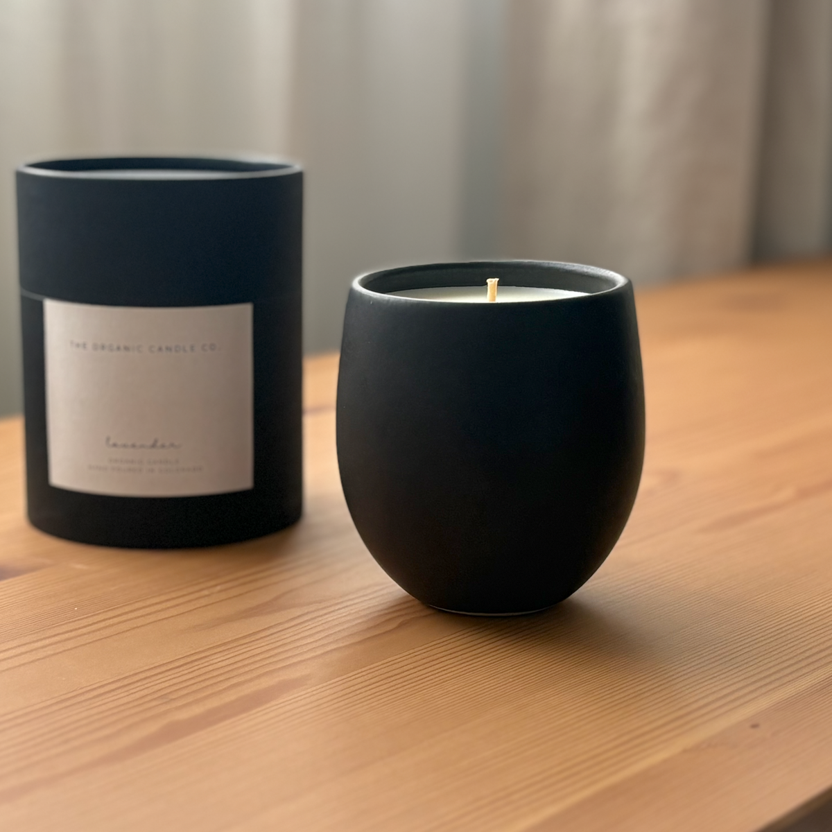 A matte black organic candle sits on a wooden table next to its matching black cylindrical packaging box, which features a white label.