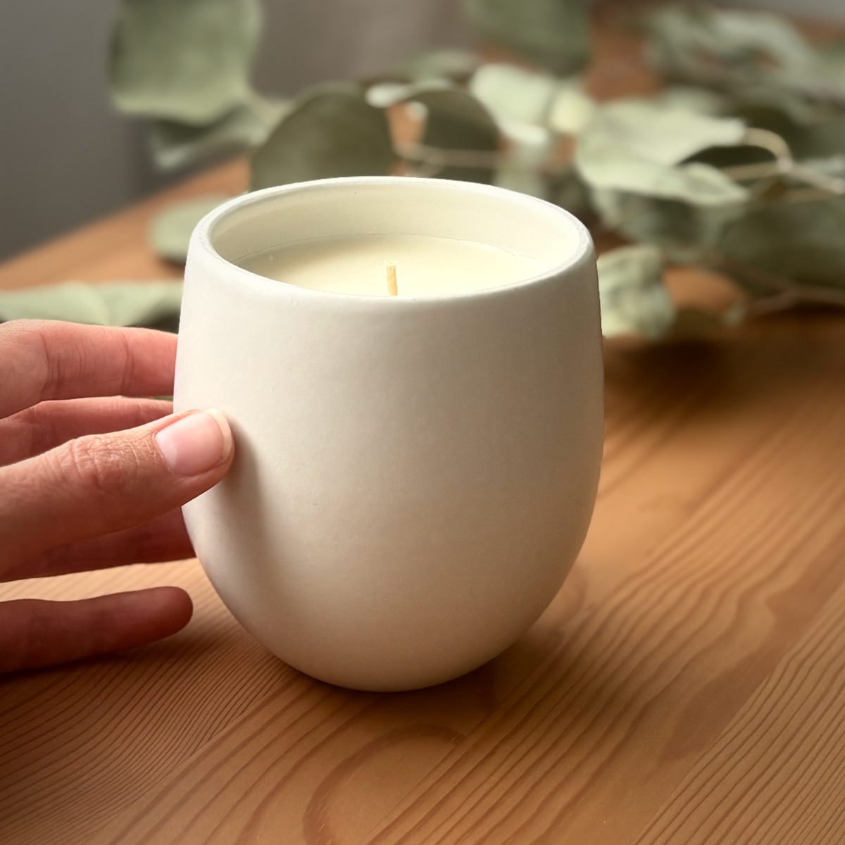 A hand holding a matte white ceramic organic candle, set on a wooden table with blurred green leaves in the background.