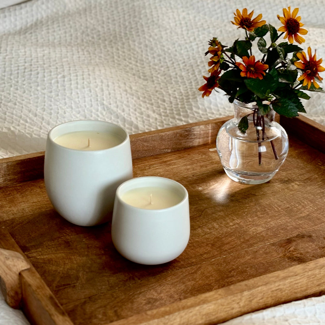 Two white ceramic organic candles on a wooden tray, with a small glass vase of orange flowers beside them, set on a textured cream fabric background.
