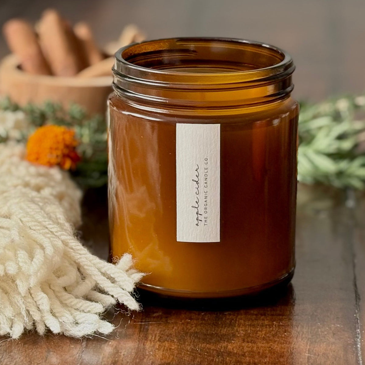 An organic candle in an amber glass jar without a lid, with a white label on a wooden surface. A blanket, cinnamon and greenery are visible to the side and background.